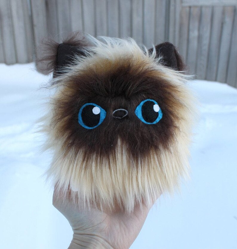 May include: A fluffy brown and white plush toy cat with blue eyes. The cat has a long, shaggy coat and a small, black nose.