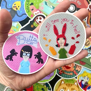 Puede incluir: Un conjunto de cuatro parches redondos bordados para planchar con personajes de la serie de televisión animada Bob's Burgers. Los parches son de color rosa, gris y azul con lunares blancos. El texto en los parches dice "Butts", "I Will See You In Hell" y "You Still Would".