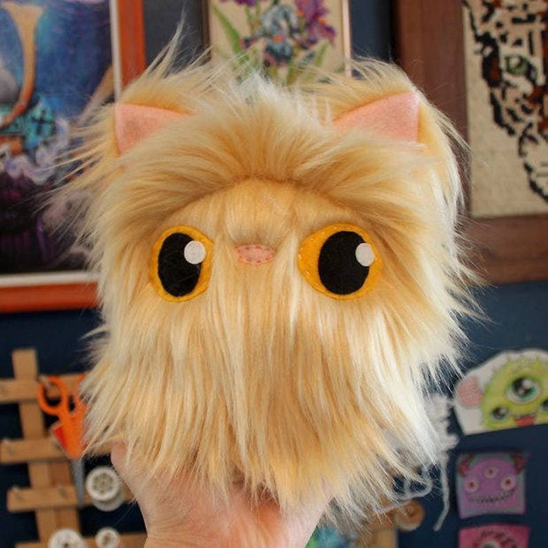 May include: A fluffy, golden-brown plush toy shaped like a cat with large, black eyes and yellow eyelids. The toy has a pink nose and a small, pink mouth.