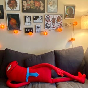 Puede incluir: Una gran figura de peluche roja con una corbata azul est&aacute; tumbada en un sof&aacute; gris. La figura tiene un ojo blanco y brazos y piernas largos. Una guirnalda de luces de calabaza naranja cuelga de la pared detr&aacute;s del sof&aacute;, que est&aacute; decorado con arte con tem&aacute;tica de Halloween.