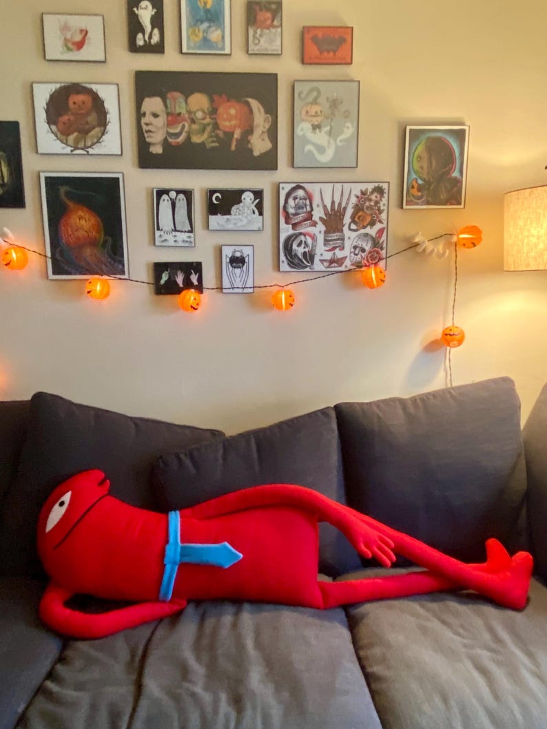 Puede incluir: Una gran figura de peluche roja con una corbata azul est&aacute; tumbada en un sof&aacute; gris. La figura tiene un ojo blanco y brazos y piernas largos. Una guirnalda de luces de calabaza naranja cuelga de la pared detr&aacute;s del sof&aacute;, que est&aacute; decorado con arte con tem&aacute;tica de Halloween.