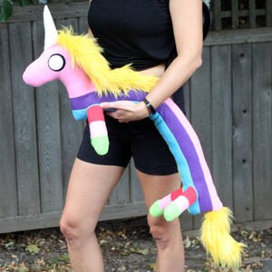 May include: A plush unicorn toy with a pink body, white horn, and yellow mane and tail. The unicorn has a rainbow-striped body and legs with green and red accents. The toy is being held by a person.