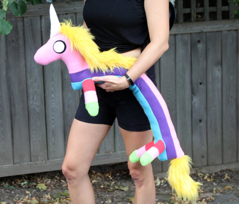 May include: A plush unicorn toy with a pink body, white horn, and yellow mane and tail. The unicorn has a rainbow-striped body and legs with green and red accents. The toy is being held by a person.