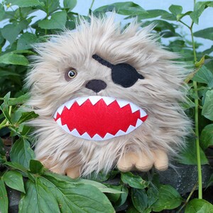 May include: A plush toy with brown fur, a red mouth with white teeth, and a black eye patch. The toy is sitting in front of green plants.