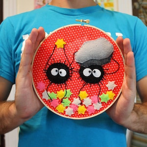 May include: A red embroidery hoop with a white polka dot pattern. Two black and white felt creatures with large eyes are holding a grey felt cloud. The creatures are surrounded by yellow, pink and green felt stars.