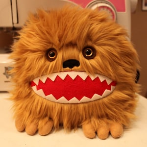 May include: A brown furry plush toy with a large, toothy grin. The toy has a red mouth with white teeth and large, round, brown eyes.