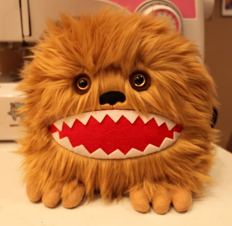 May include: A brown furry plush toy with a large, toothy grin. The toy has a red mouth with white teeth and large, round, brown eyes.