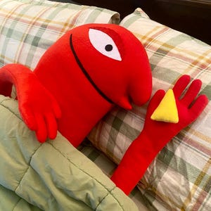 Puede incluir: Un personaje de peluche rojo con un ojo blanco y una sonrisa negra, sosteniendo un tri&aacute;ngulo amarillo. El personaje est&aacute; en la cama, cubierto por una manta verde, con almohadas a cuadros en el fondo.