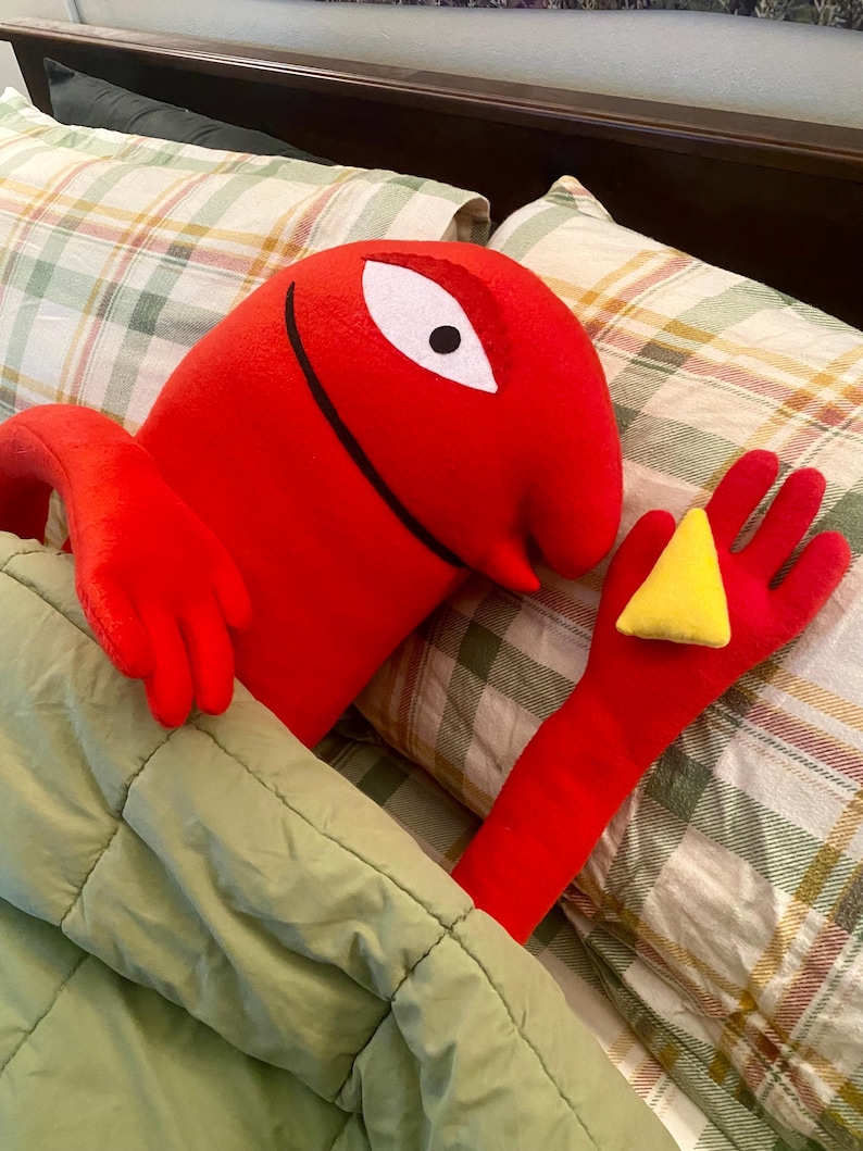 Puede incluir: Un personaje de peluche rojo con un ojo blanco y una sonrisa negra, sosteniendo un tri&aacute;ngulo amarillo. El personaje est&aacute; en la cama, cubierto por una manta verde, con almohadas a cuadros en el fondo.