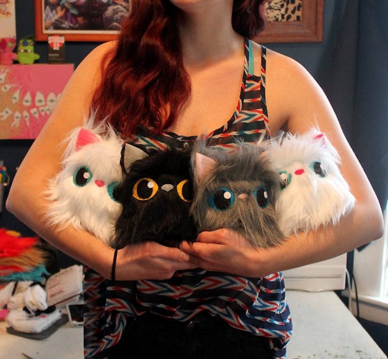 May include: A woman is holding four plush toys. The toys are shaped like cats and are white, black, and gray. The woman is wearing a striped tank top.