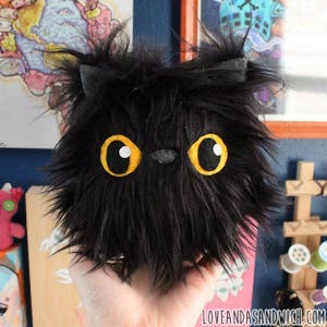 May include: A black plush toy cat with large yellow eyes and a black nose. The cat is made of soft, fluffy material and has a round, cuddly shape.