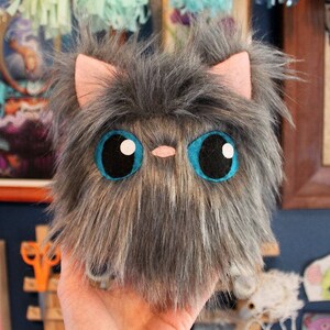 May include: A plush gray and brown cat toy with large blue eyes and a pink nose. The toy is made of soft, fluffy material and is perfect for cuddling.