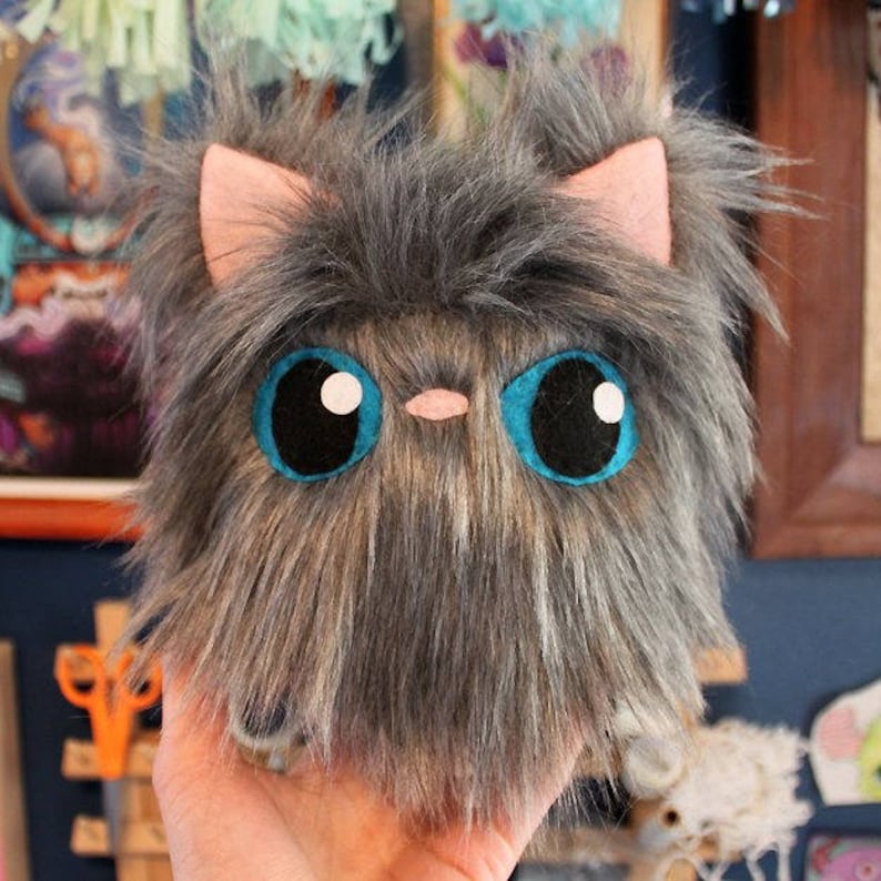 May include: A plush gray and brown cat toy with large blue eyes and a pink nose. The toy is made of soft, fluffy material and is perfect for cuddling.