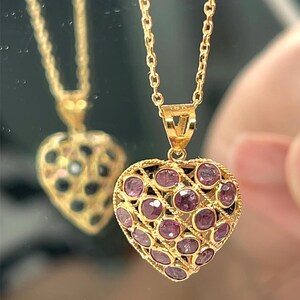 May include: A gold heart pendant necklace. The pendant is filled with pink gemstones and has an openwork design. The necklace chain is also gold. The pendant is reflected in a mirror.