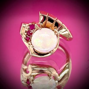 May include: A gold ring with a large, round, opalescent stone at its centre, complemented by small red gemstones. The ring has an ornate, sculpted design with a polished finish. The ring is displayed on a reflective pink surface.