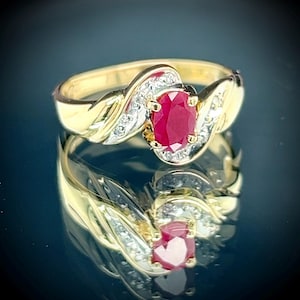May include: A gold ring with a large oval ruby gemstone at its centre. The ruby is a deep red colour and is surrounded by small, sparkling diamonds. The ring has a twisted, wave-like design.