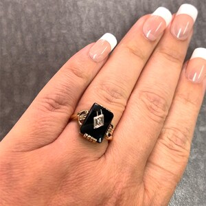 May include: A gold ring with a rectangular black onyx stone and a small diamond. The ring has detailed gold work on the band and around the onyx. The ring is displayed on a hand with manicured nails.