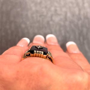 May include: A gold ring featuring a rectangular black onyx stone and a small diamond. The ring has a detailed, vintage design with a decorative band.