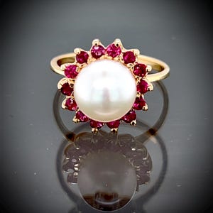 May include: A gold ring featuring a large, round white pearl at the center, surrounded by a halo of small, vibrant red gemstones. The ring's band is a simple, polished gold. The ring is displayed on a reflective surface.