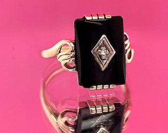 May include: A gold ring featuring a rectangular black onyx stone with a diamond-shaped accent. The ring has a decorative gold band with ornate detailing on the sides. The ring is displayed on a reflective surface.