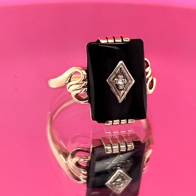 May include: A gold ring with a rectangular black onyx stone and a diamond-shaped detail. The ring has a decorative gold band with ornate side detailing. The ring is shown on a reflective surface.