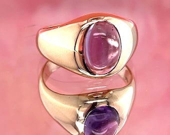 May include: A gold ring with a large, oval, pink gemstone. The ring is set in a polished gold band, and the gemstone has a smooth, rounded surface. The ring is displayed on a reflective pink surface.