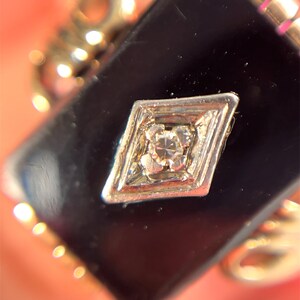 May include: Close-up of a vintage pendant with a black onyx stone. A small, diamond-shaped silver setting with a central diamond is set on the onyx. The setting has a diamond-shaped design.