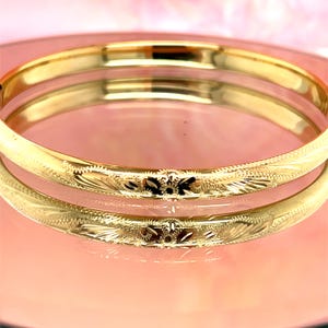 May include: A gold-colored bangle bracelet with an engraved floral design. The bracelet is reflected in a pink surface, creating a mirrored effect. The intricate details of the engraving are visible.