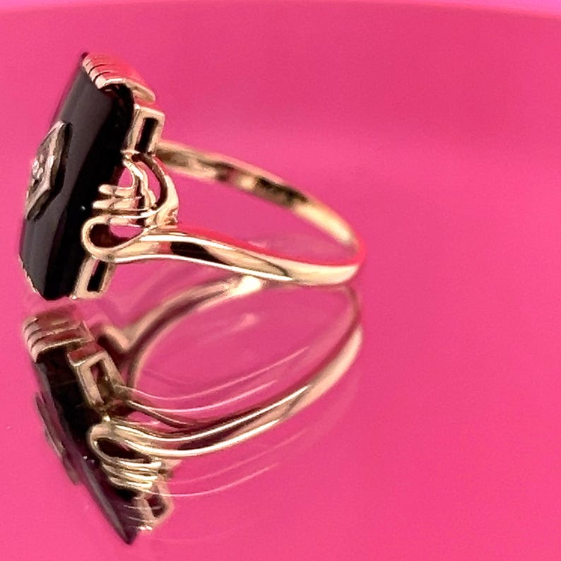 May include: A gold ring featuring a black onyx stone and a small diamond. The ring has an Art Deco design with a rectangular onyx stone and detailed gold filigree. The ring is set against a pink background.