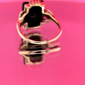 May include: A gold ring featuring a rectangular black gemstone. The ring has an ornate setting and a curved band. The ring is reflected on a pink surface.