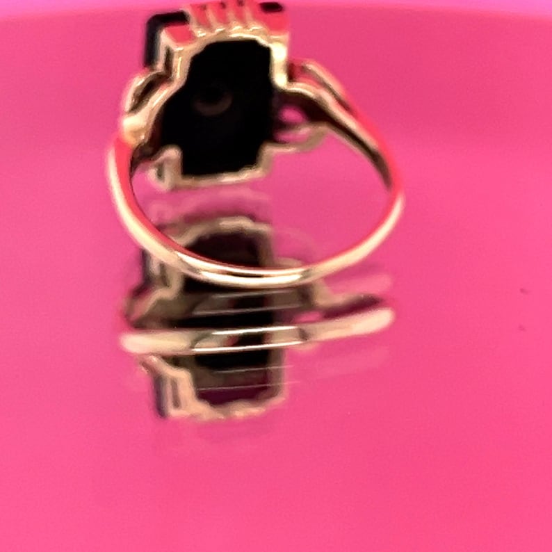 May include: A gold ring featuring a rectangular black gemstone. The ring has an ornate setting and a curved band. The ring is reflected on a pink surface.