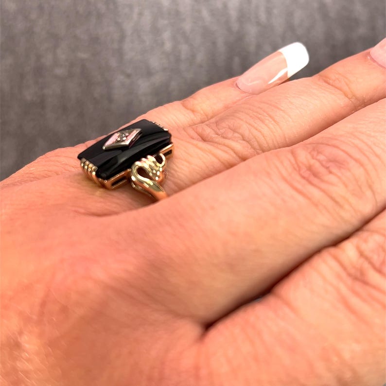 May include: A gold ring with a rectangular black onyx stone and a diamond-shaped accent. The ring has an ornate gold setting with decorative details. The ring is on a finger.