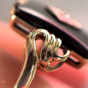 May include: Close-up of a gold ring with an ornate design. The ring features a dark, rectangular stone. The gold setting has intricate details, with a curved, looped design. The background is a soft, blurred pink.
