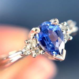 May include: A detailed shot of a silver ring with a prominent, pear-cut, blue gemstone. The ring is embellished with small, clear accent stones. The band is slender and polished. The ring's design is elegant and refined.