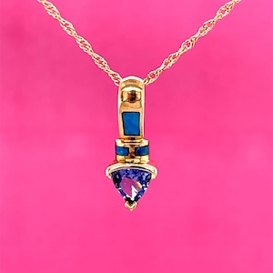 May include: A gold pendant necklace featuring a blue and purple gemstone. The pendant has a rectangular blue stone and a triangular purple stone. The chain is a twisted rope design, set against a pink backdrop.