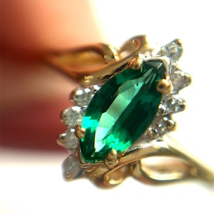 May include: A gold ring featuring a central marquise-cut green gemstone surrounded by a cluster of small, clear, round-cut accent stones. The metal band has a polished finish and a delicate, curved design that supports the gemstone setting.