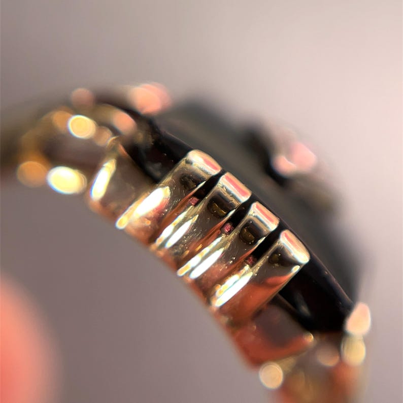 May include: Close-up of a gold ring featuring a series of rectangular, ridged details. The ring has a dark, reflective band. The image is in soft focus, with blurred highlights.