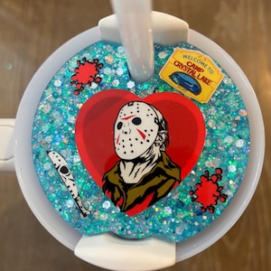 Puede incluir: Una taza blanca y azul con una pajita, con un diseño azul brillante. El diseño incluye un corazón rojo con una máscara de Jason Voorhees, un cuchillo y el texto "WELCOME TO CAMP CRYSTAL LAKE".