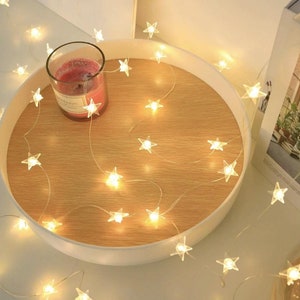 Può includere: Un vassoio rotondo in legno con bordo bianco contiene una candela accesa in un barattolo di vetro. Una stringa di piccole luci a forma di stella è drappeggiata sul vassoio. Lo sfondo include una mensola bianca e una stampa incorniciata.