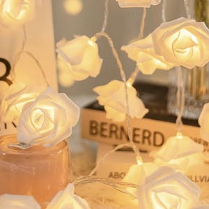 May include: A decorative string of white rose fairy lights. The illuminated roses emit a warm glow. The lights are strung on a thin, clear wire. The background features a blurred book and a candle.
