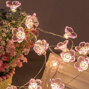 Puede incluir: Una cadena de luces de hadas con bombillas en forma de flores rosas. Las luces están enhebradas en un alambre delgado y son perfectas para agregar un toque de fantasía a cualquier espacio.