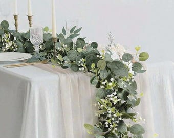 May include: A white tablecloth with a eucalyptus garland with white flowers running down the center. The garland is on a table with candlesticks and a glass vase.