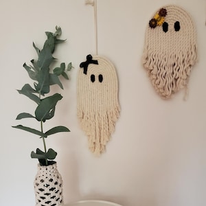 Könnte beinhalten: Eine weiße Keramik-Geisterfigur steht auf einem weißen Tisch neben einer Glasflasche, die mit einem Makramee-Design umwickelt ist. Zwei Makramee-Geister-Wandbehänge mit schwarzen Augen und Schleifen sind ebenfalls zu sehen.