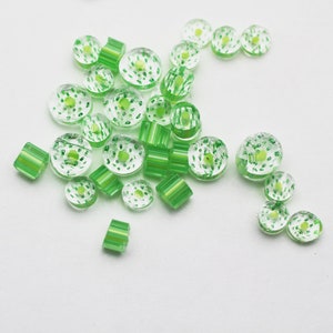 May include: A collection of green glass beads with white speckles. Some beads are round, while others are cylindrical. The beads are scattered on a white surface.