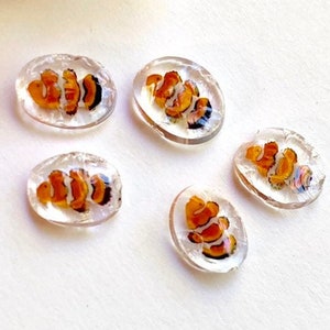May include: Five small, oval-shaped glass beads with a clear background and an orange and black fish design inside.
