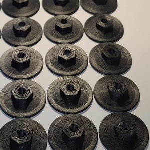 Op de afbeelding: Een verzameling van 12 zwarte plastic schroefdoppen met een zeskantig midden. De doppen zijn in rijen op een wit oppervlak gerangschikt.