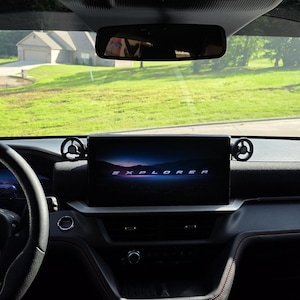 May include: The interior of a vehicle featuring a black steering wheel with a Ford logo, a central infotainment screen displaying the word EXPLORER in white text against a dark landscape background, and two circular air vents on the dashboard.