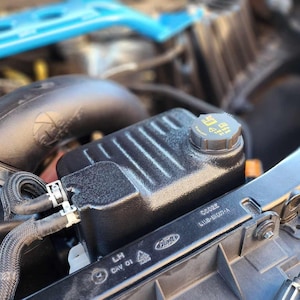 Könnte beinhalten: Nahaufnahme eines schwarzen Motorraums mit einem schwarzen Kunststoffbehälter mit gelbem Deckel mit der Aufschrift "5.0L AT", einem schwarzen Schlauch mit einer silbernen Klemme und einem schwarzen Lufteinlass mit dem Text "GT MENTUM" darauf.