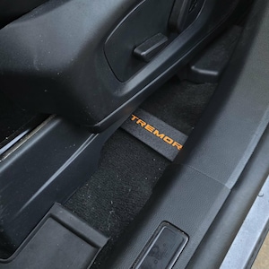 May include: Interior view of a vehicle's front seat area. The image shows a black seat, floor mat, and door panel. The floor mat has the word "TREMOR" in orange lettering.