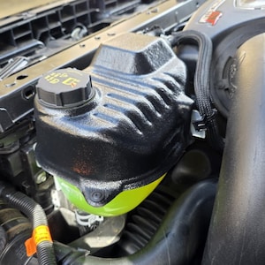 May include: Close-up of a car engine compartment. The image highlights a black coolant reservoir with a textured surface and a black cap. The reservoir contains a bright green liquid. Various engine components and hoses are visible.